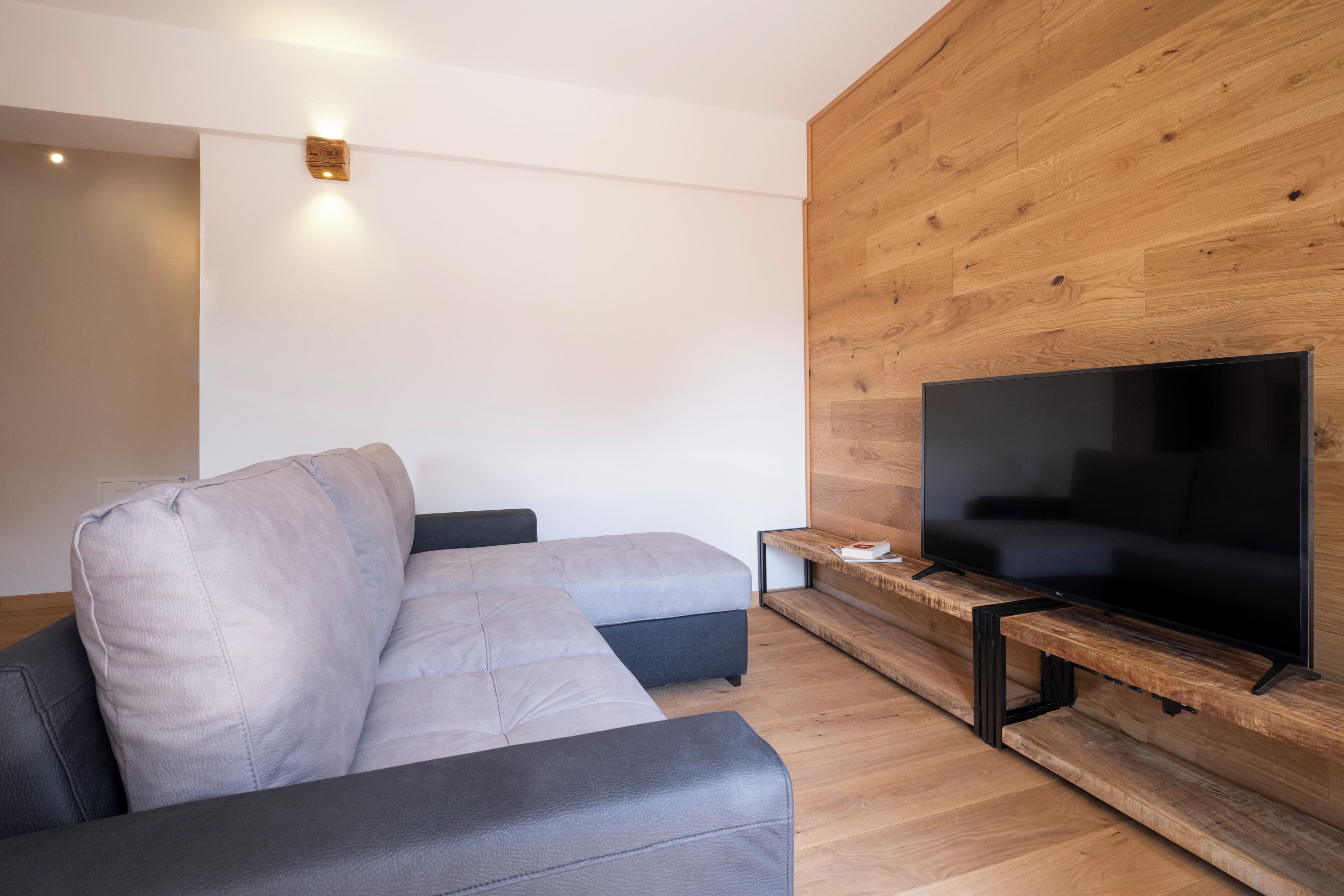 A modern living room with a grey sectional sofa, a flat-screen TV mounted on a wooden wall, and a small wooden side table.