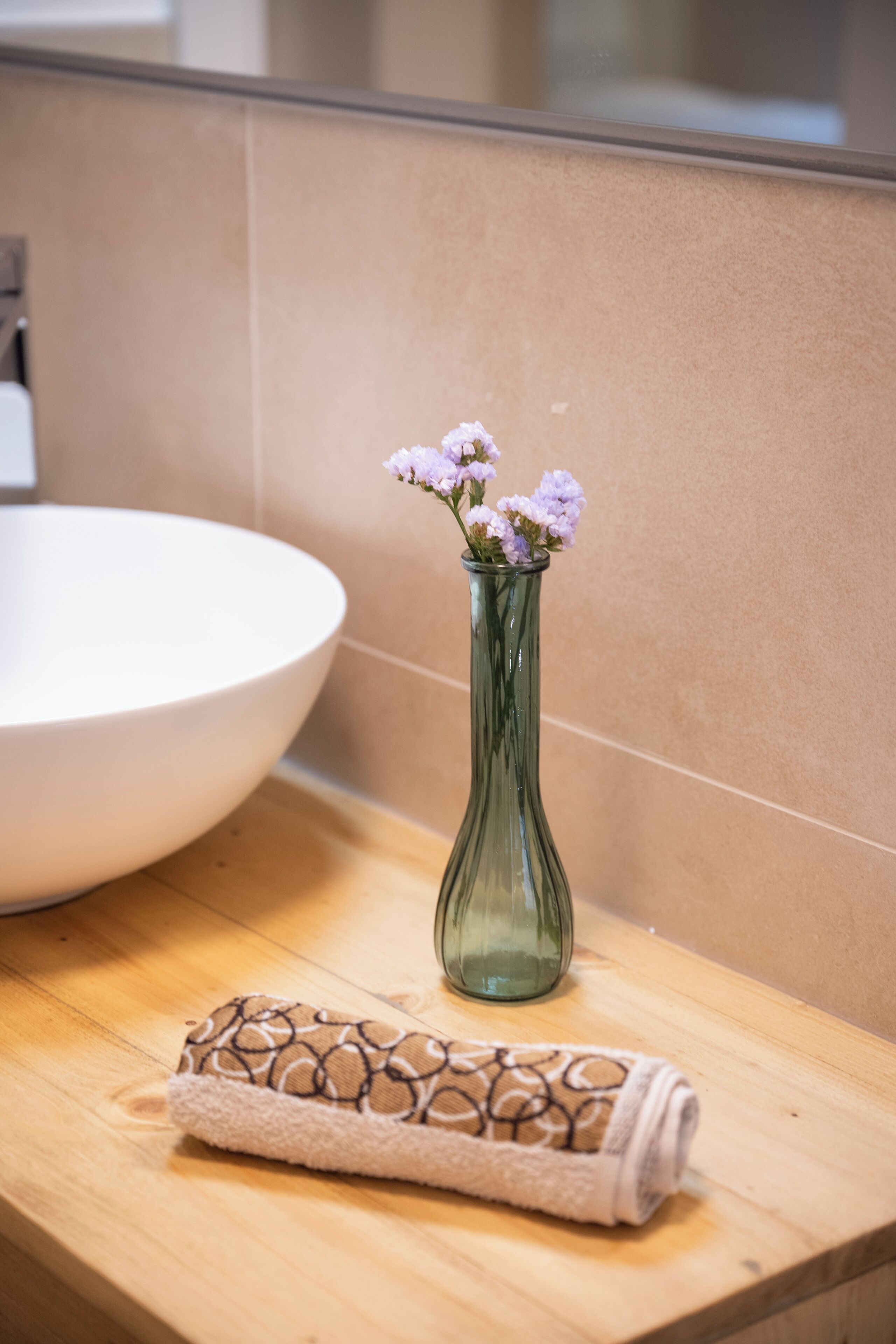 A bathroom with a large white basin, a green vase with flowers, and a rolled towel on a wooden countertop.