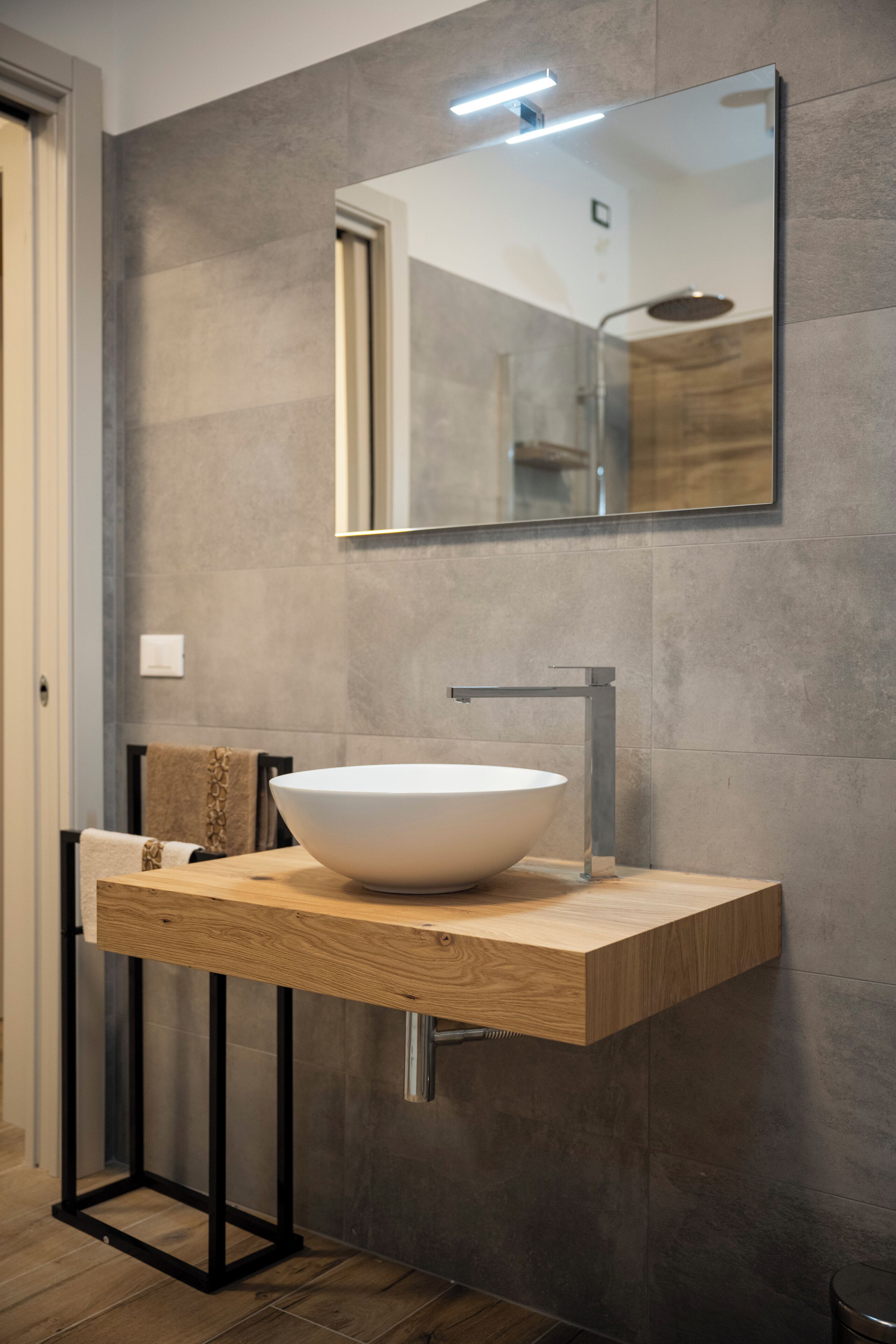 Un baño moderno con un lavabo blanco de diseño, un mueble de tocador de madera, un soporte de metal negro y un espejo sobre el lavabo.