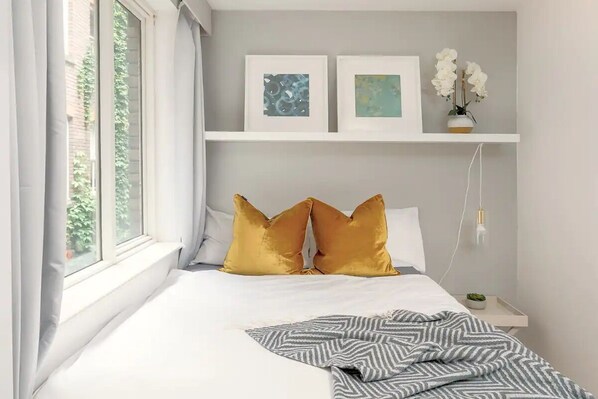 A bedroom with a bed, two gold pillows, a gray and white striped blanket, a nightstand with a small plant, and two framed artworks on the wall. - Spacious 2 bedroom in Liverpool Street (London)