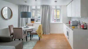 A modern kitchen with white cabinets, a washing machine, and a microwave. There is a dining area with a round table and chairs, and a large mirror on the wall. - Spacious 2 bedroom in Liverpool Street (London)
