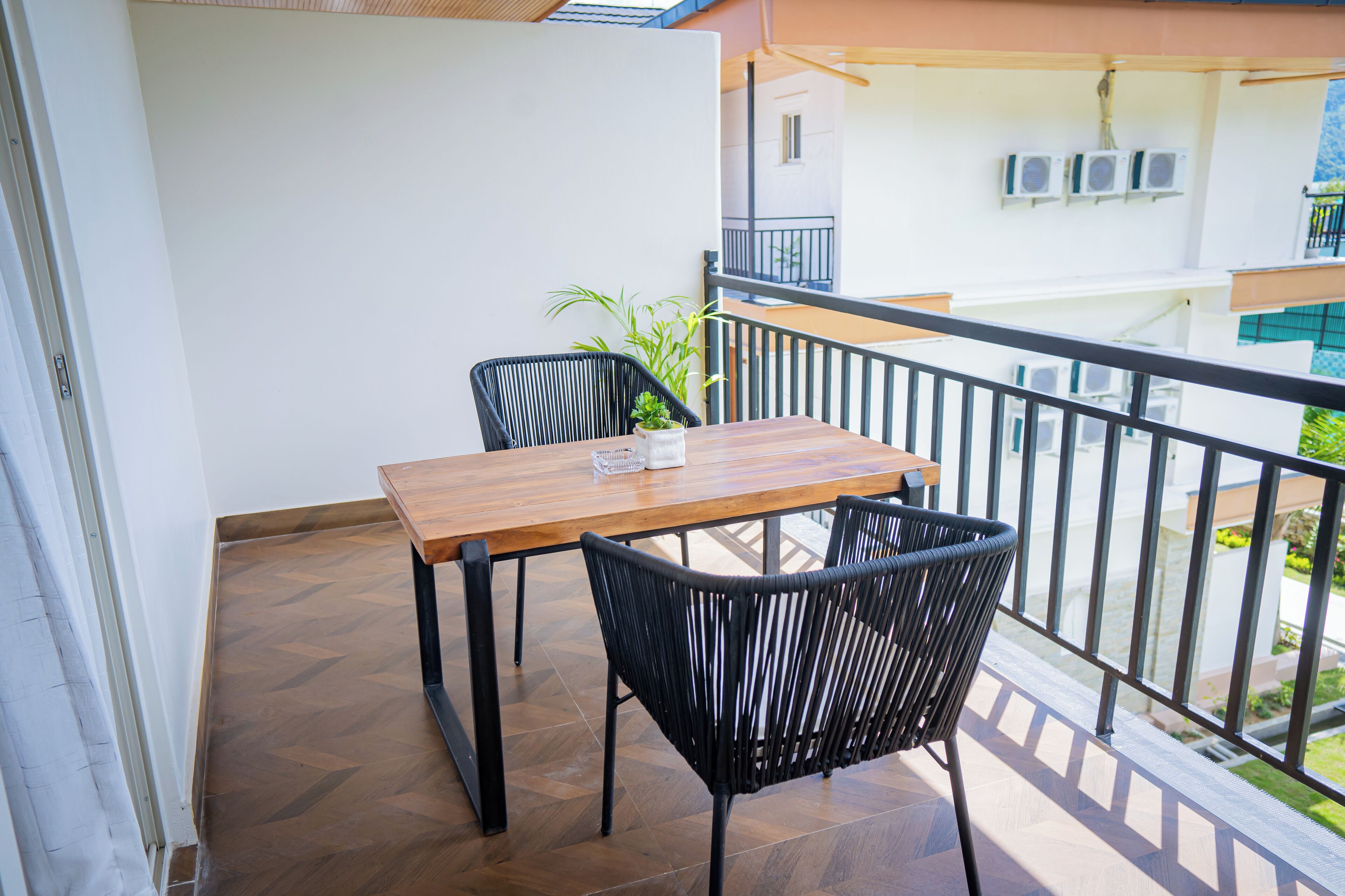 A balcony with a wooden table and two black chairs, a potted plant, and a view of a building with air conditioning units.