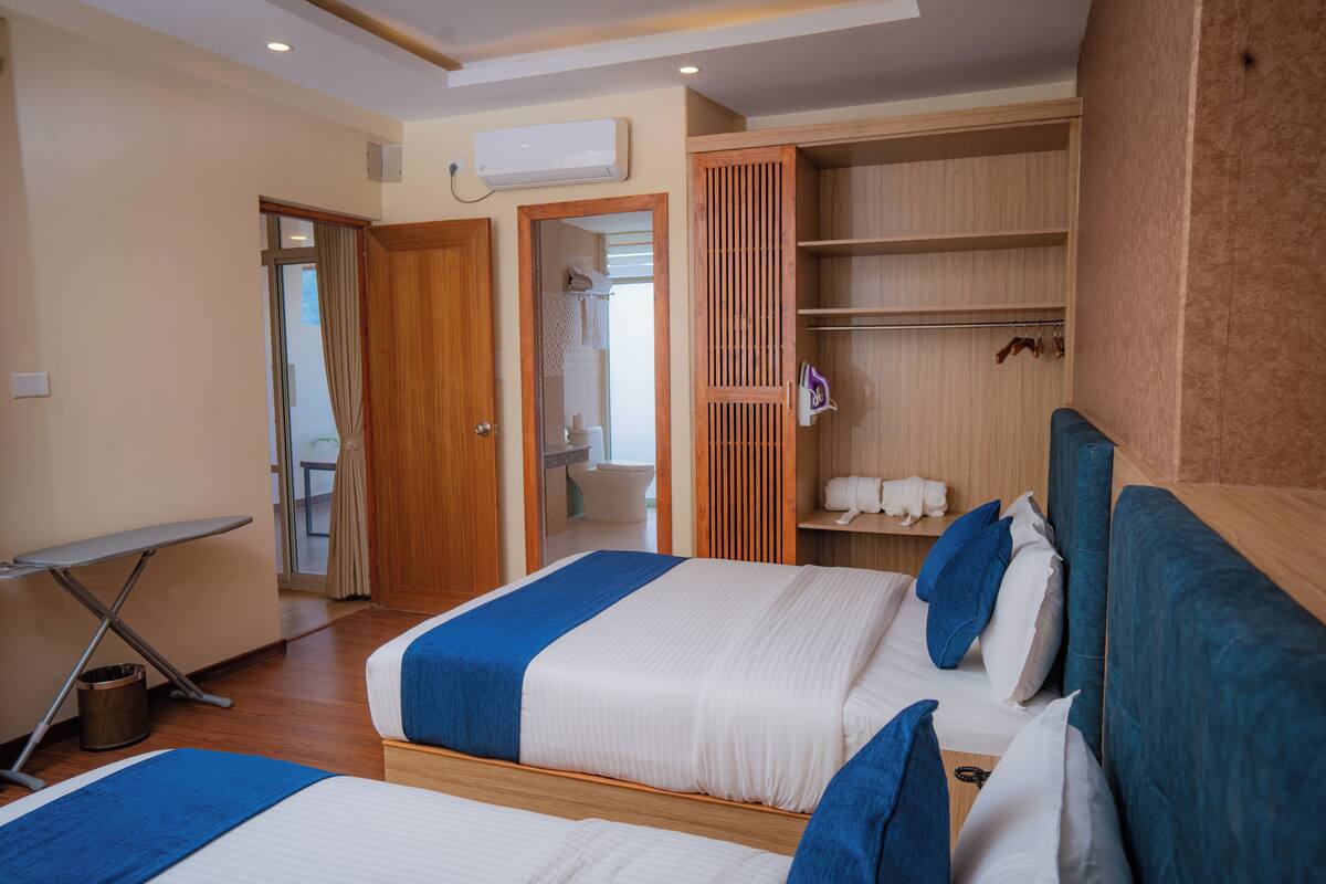 Ein Hotelzimmer mit zwei Betten, einem Holzschrank, einem durchgehenden Türspalt erkennbaren Badezimmer und einem kleinen Tisch mit Bügelbrett.