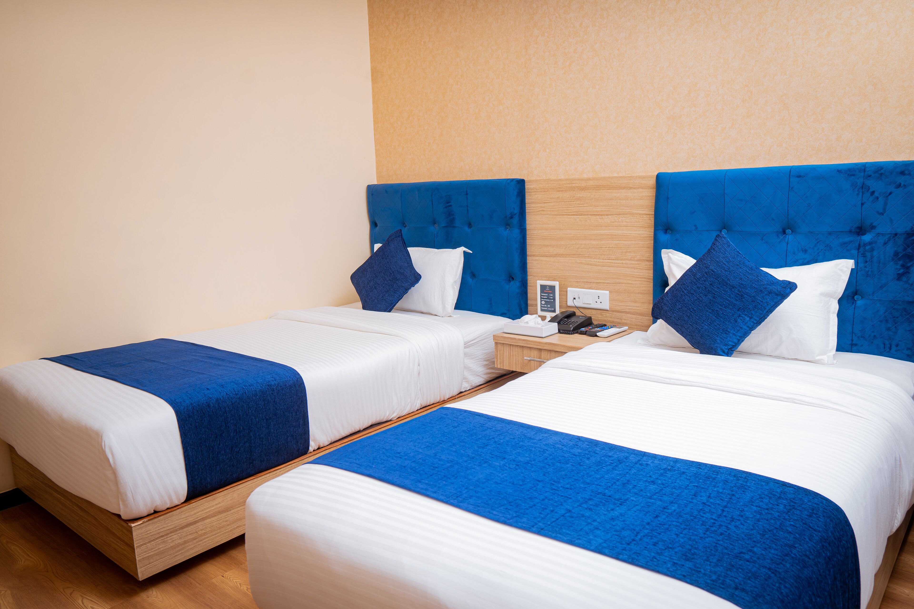 A hotel room with two beds, each with a blue headboard and white bedding. There is a wooden nightstand with a phone and a small table between the beds.