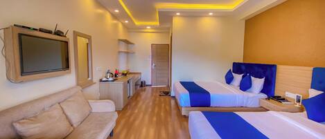 A hotel room with a sofa, two beds with blue and white bedding, a TV mounted on the wall, and a small kitchenette area.