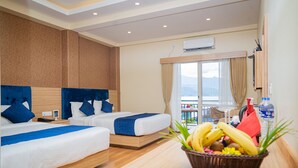 Habitación de hotel con dos camas, cabecero de madera, ventana con cortinas, balcón y una cesta de frutas sobre la mesa.