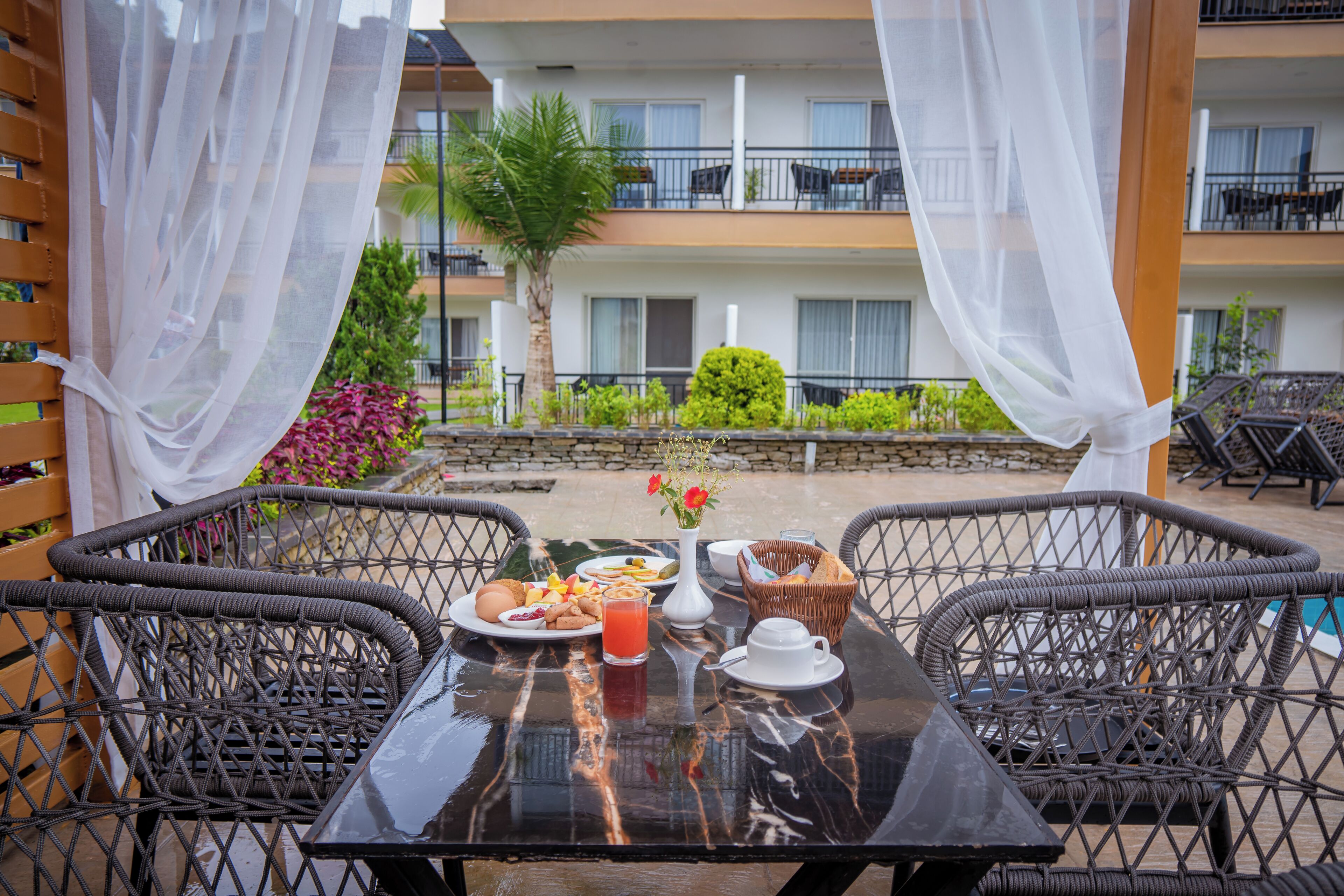 A dining table with wicker chairs, a breakfast setting, and a view of a building with balconies and greenery.