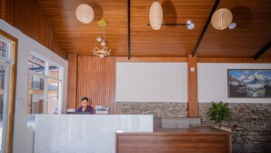 Mostrador de recepción con recepcionista, techo de madera, pared de piedra y una pintura de montañas.