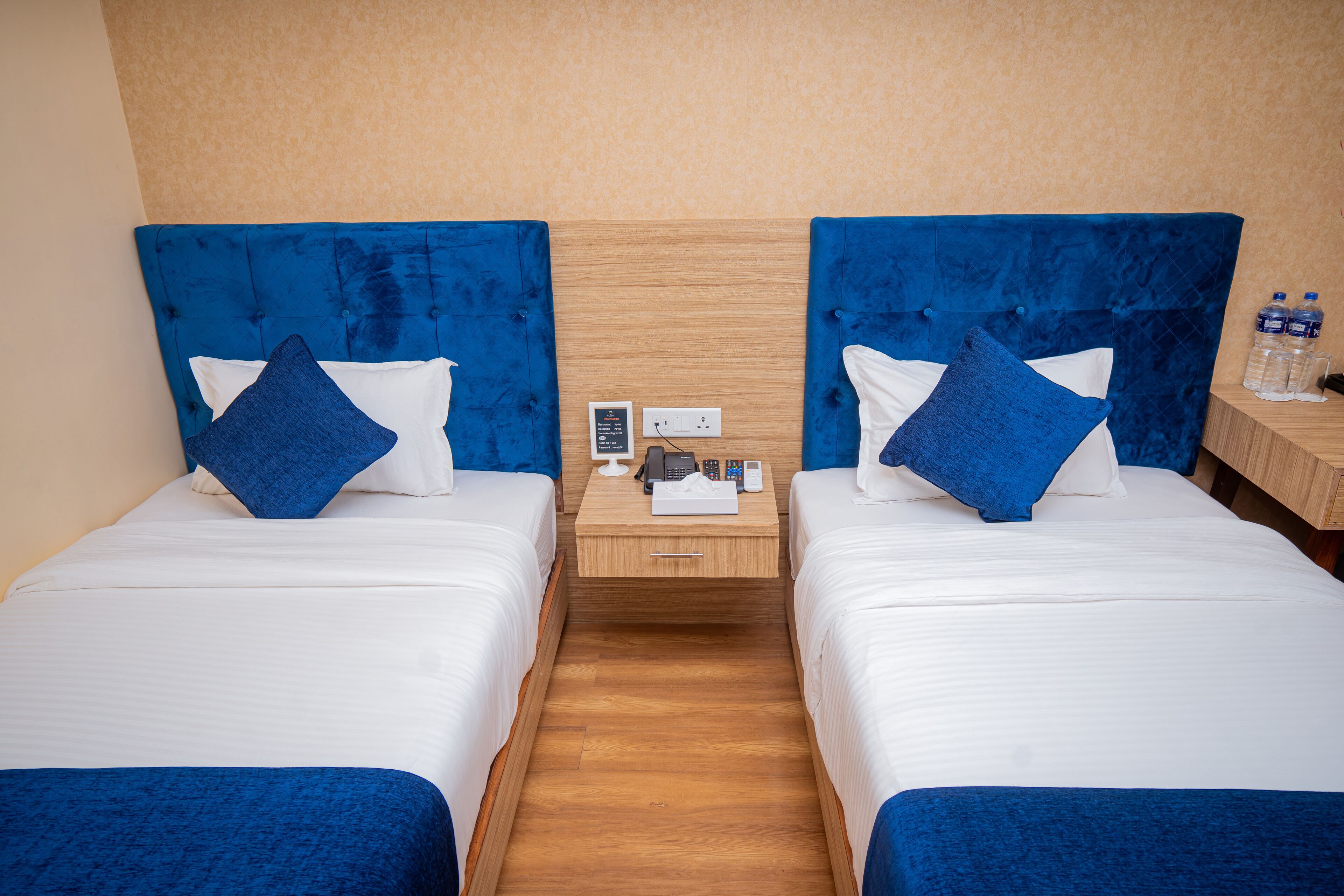 A hotel room with two beds, each with a blue headboard and white bedding. There is a wooden nightstand between the beds with a phone and a small table with a lamp.
