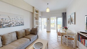 A living room with a beige sofa, a dining table with chairs, a bookshelf, and a view of the sea through the open door. - Aneria Suites (Rethymno)
