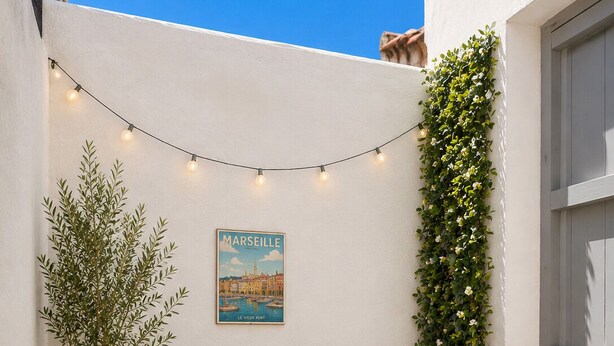 A small outdoor patio with a table, chairs, a potted plant, a wall-mounted poster, and string lights.