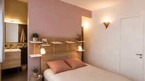 A bedroom with a wooden headboard, bedside tables, a bed with pillows, a nightstand, a wall-mounted shelf with plants, and a door.