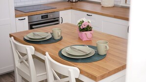 Une cuisine avec des meubles blancs, un plan de travail en bois et une table à manger dressée avec des assiettes, une tasse et une plante en pot.