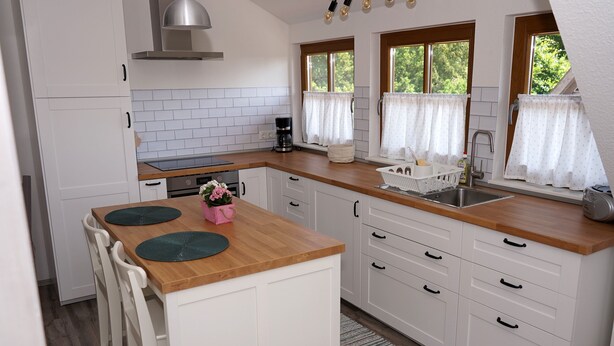 Une cuisine moderne avec des meubles blancs, un plan de travail en bois et un îlot central. Elle comprend deux fenêtres avec des rideaux, une cuisinière encastrée et une machine à café.
