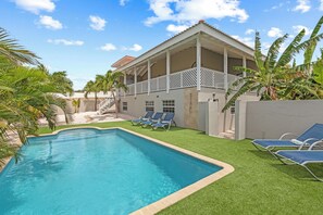 Una piscina con solárium blanco, rodeada de palmeras y un edificio con balcón.