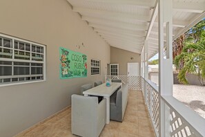Un espacio al aire libre techado con una mesa larga y bancos, una pared con un mural colorido, y vistas a palmeras.
