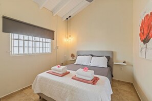 A bedroom with a bed, two bedside tables, a window with blinds, a wall-mounted light fixture, and a painting.
