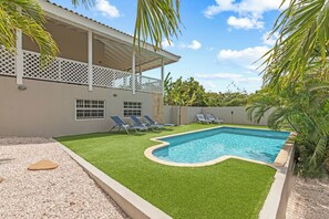 A swimming pool with a curved edge, surrounded by artificial grass, and adjacent to a building with a balcony.