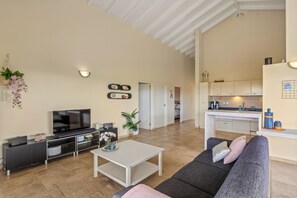 Una sala de estar moderna con televisor de pantalla plana, un sofá y una mesa de centro. Se observa un área de cocina con armarios y una encimera al fondo.