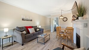 Un salon comprenant un canapé, une table basse et un coin repas avec des chaises. On y trouve une cheminée agrémentée d’un miroir et d’une plante posée sur le manteau.