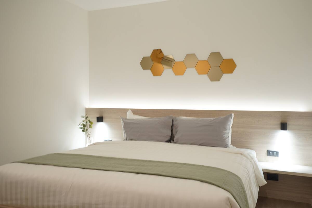 A neatly made bed with a wooden headboard, a wall-mounted hexagonal light fixture, and a bedside table with a lamp.