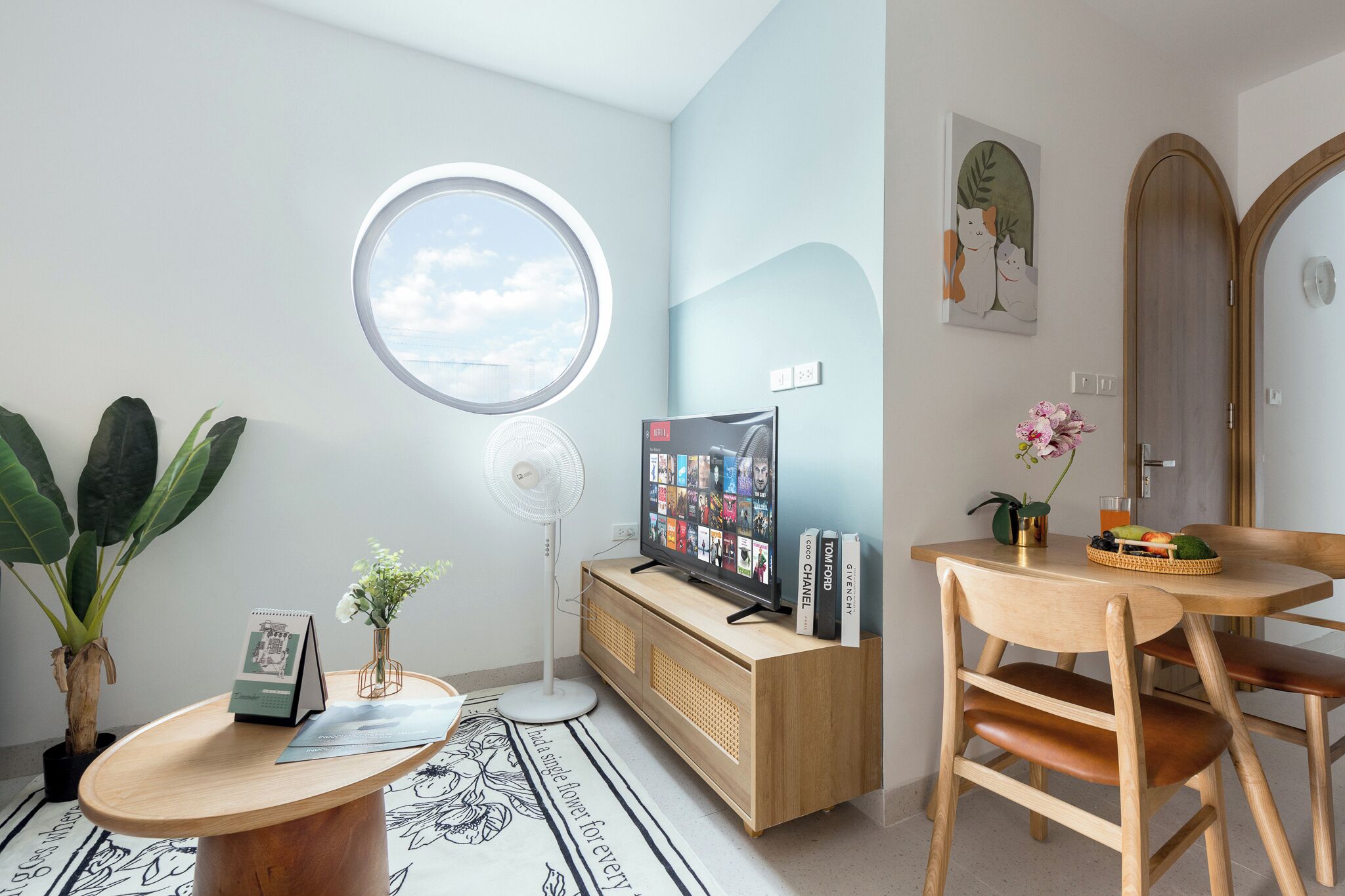 Un soggiorno moderno con una finestra circolare, una TV su una consolle di legno, un tavolo da pranzo con sedie e una pianta in vaso.