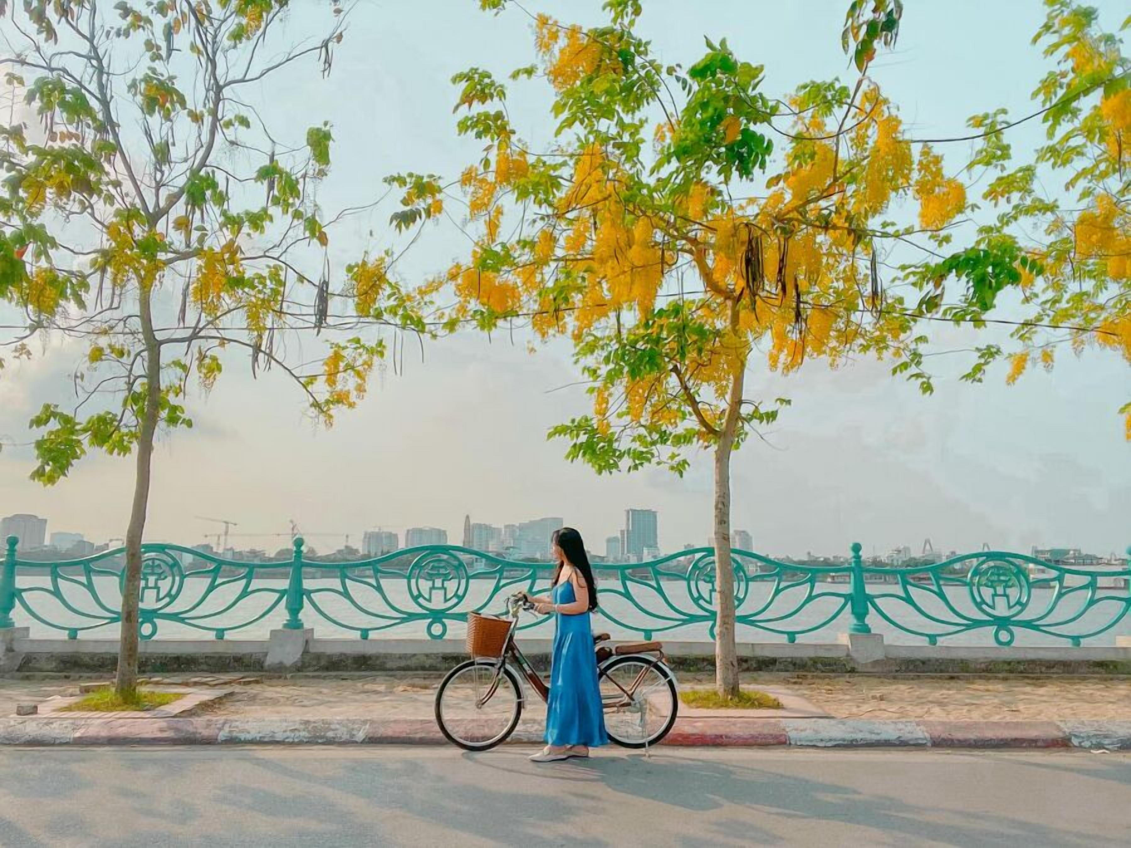 Une personne se tient à côté d’un vélo sur un trottoir, avec une clôture décorative et des arbres en arrière-plan.