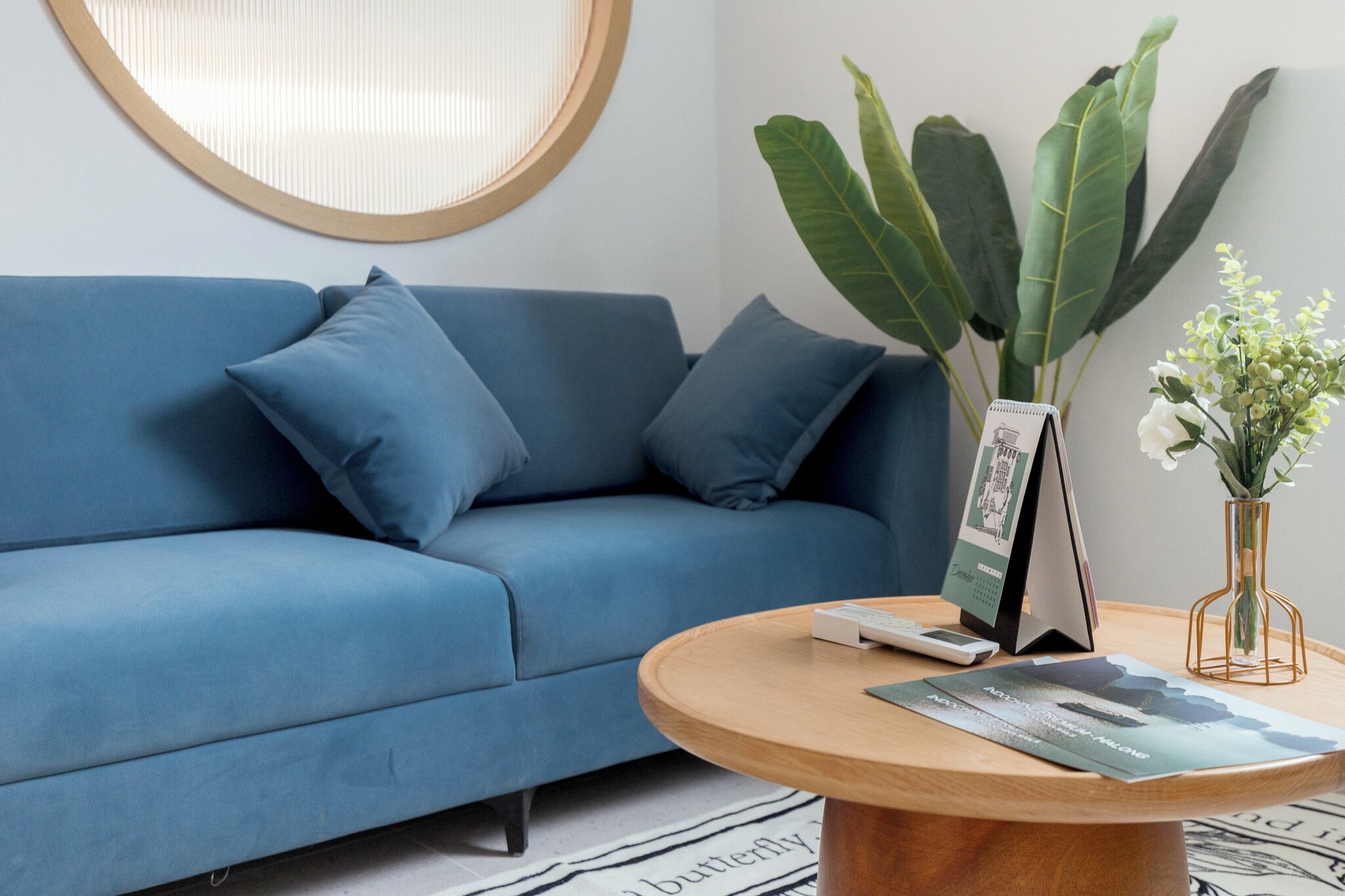 A blue sofa with matching cushions, a round wooden coffee table with a magazine, a framed picture, and a vase with white flowers.