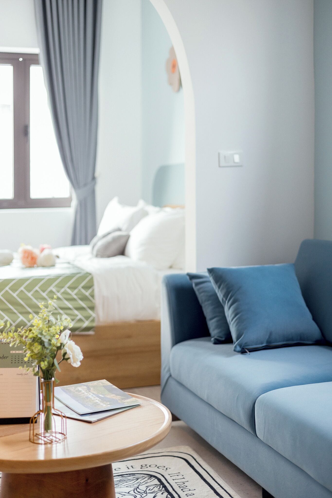 Un dormitorio moderno con cama, sofá azul y una mesa redonda de madera con un jarrón y un libro.