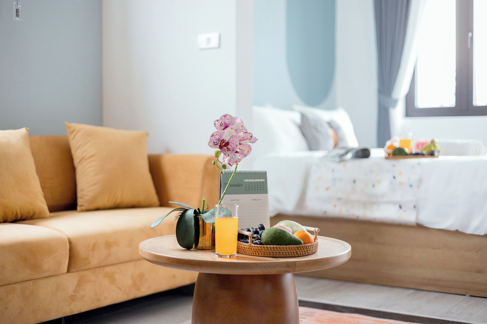 A cozy room with a sofa, a small round table with a glass of orange juice, a vase with a pink orchid, and a basket of fruits.