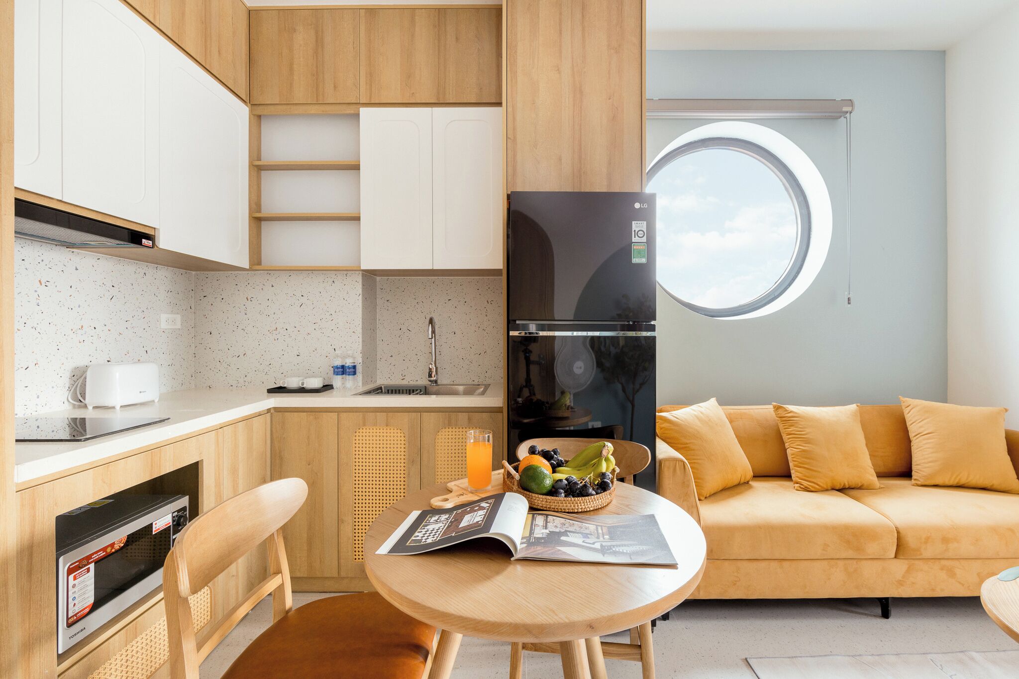 A compact kitchen with a microwave, toaster, and sink. There's a dining area with a table, chairs, and a magazine. A sofa and a window with a view are also visible.