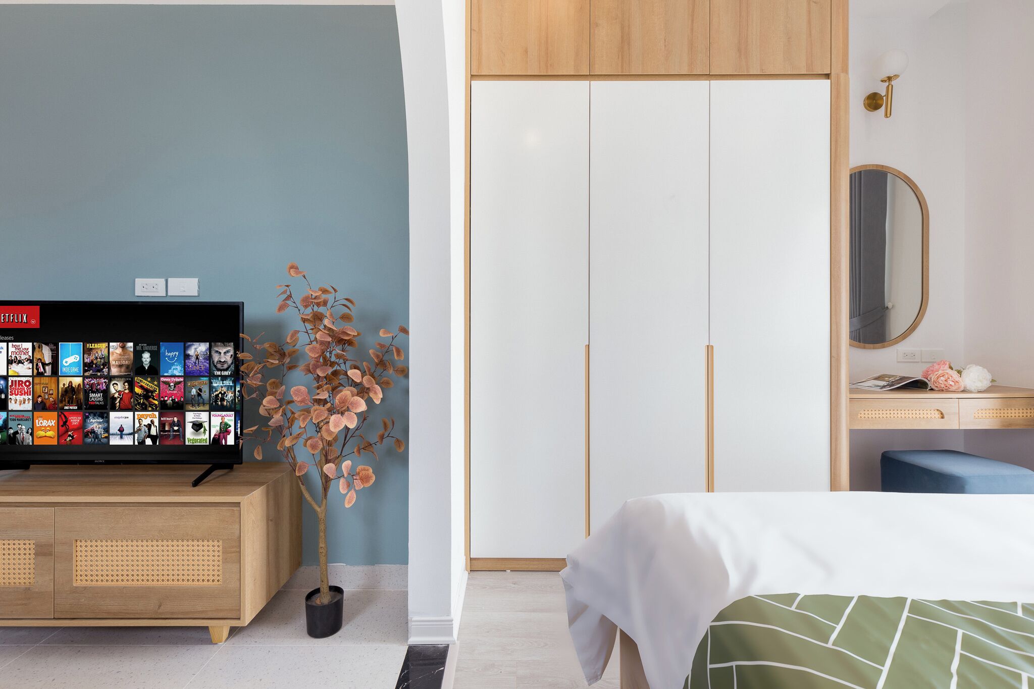 A hotel room with a TV displaying a variety of channels, a wooden cabinet, a potted plant, a wardrobe, a desk with a mirror, and a bed with a green and white bedspread.