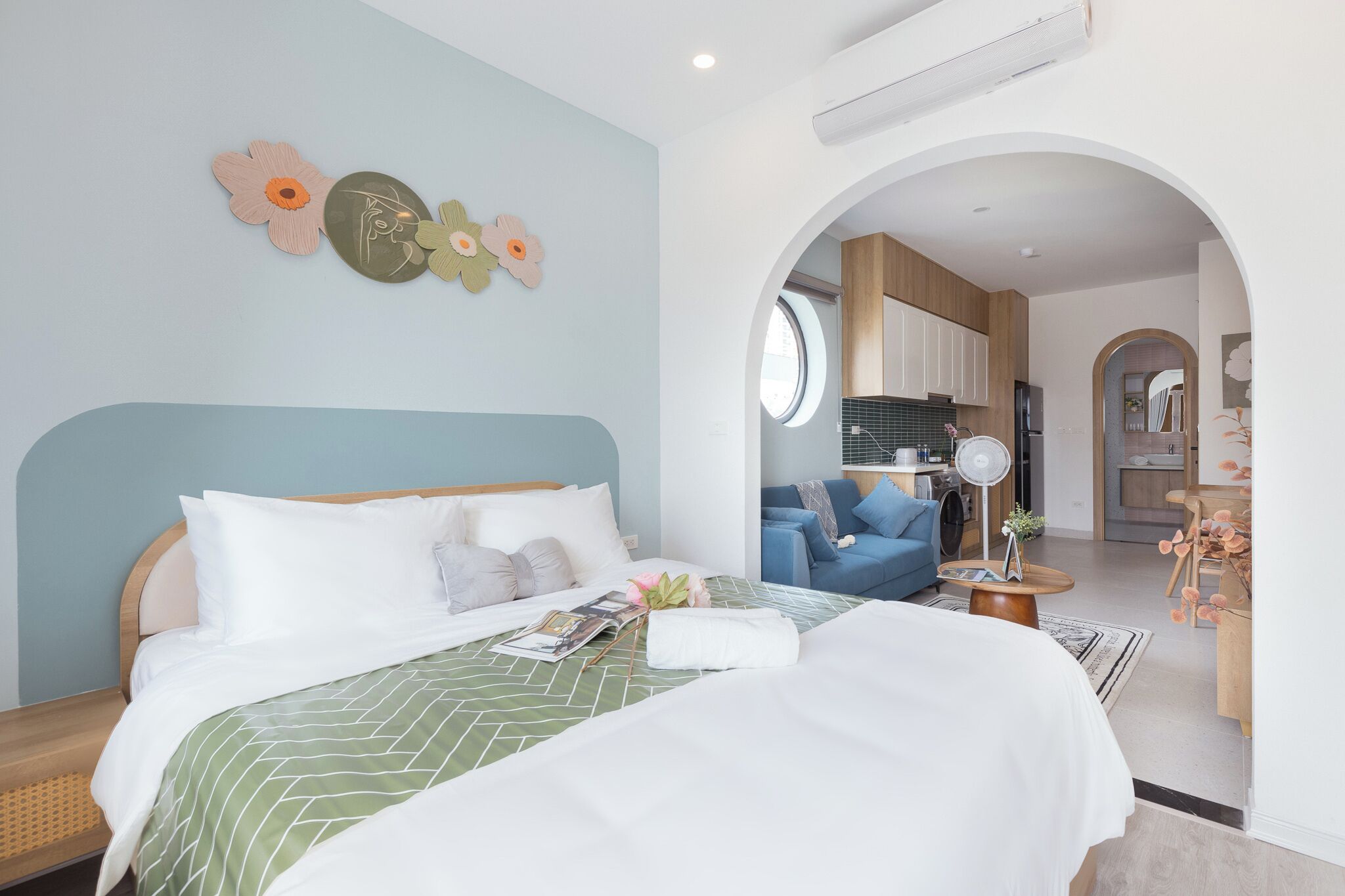 Une chambre bien éclairée, dotée d’un grand lit, d’une table de chevet et d’une commode. Une fenêtre offre une vue sur la cuisine et le salon.
