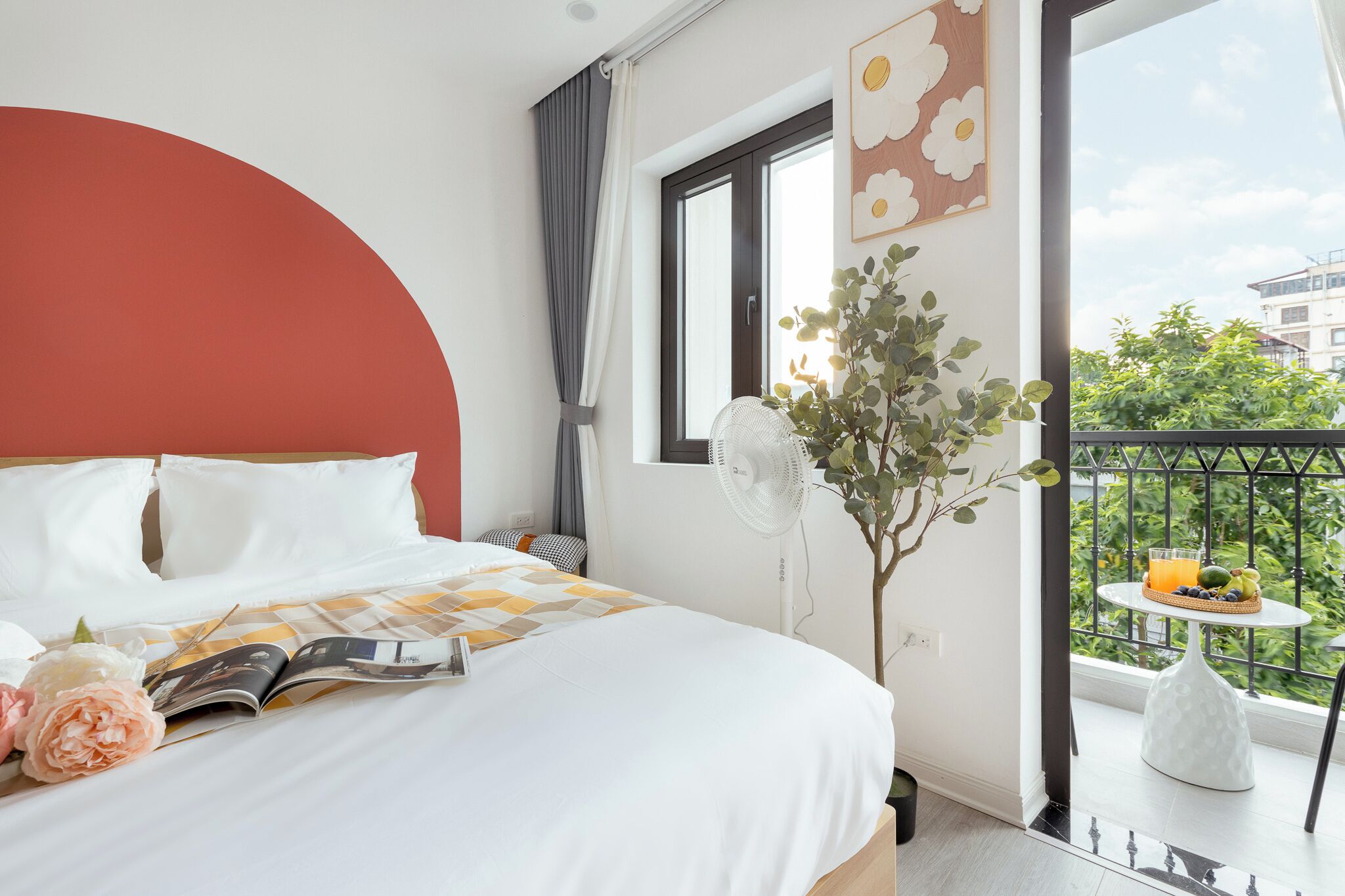 A hotel room with a bed, a window with a view of greenery, a small balcony with a table and a fan.