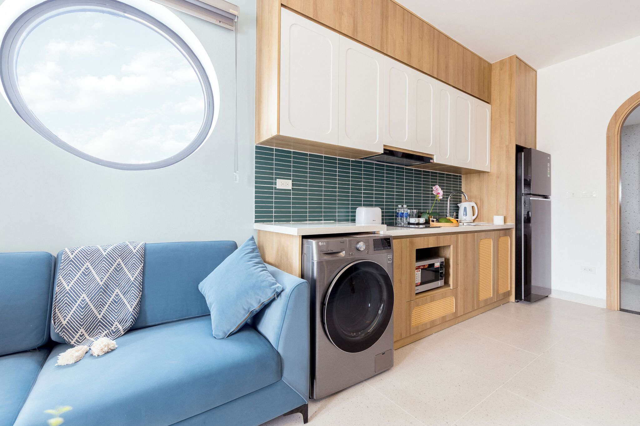 A compact kitchen with a washing machine, a dining area with a blue sofa, and a view through a window.
