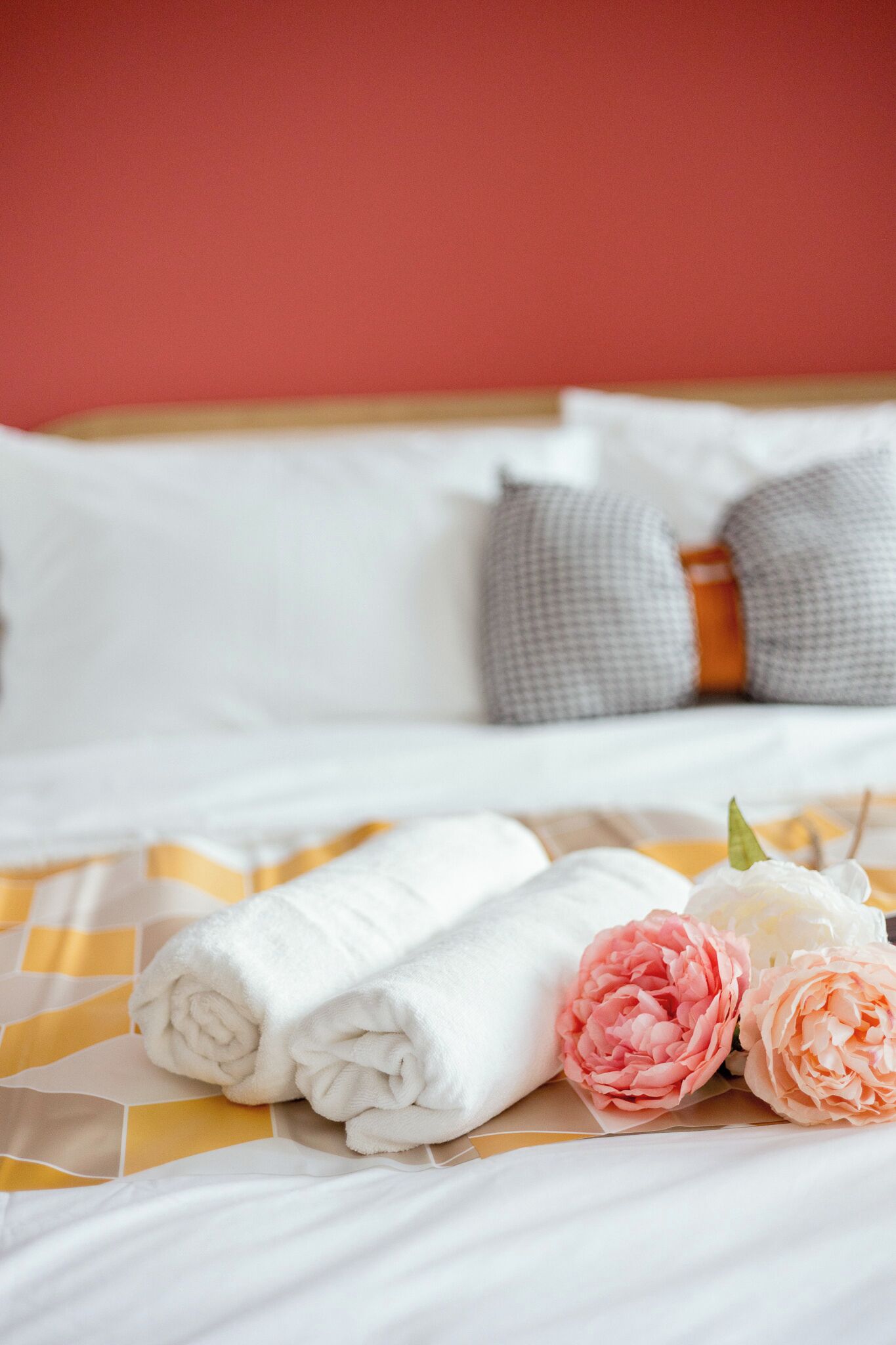 Una cama bien hecha con almohadas blancas y estampadas, una colcha de cuadros amarillos y blancos, y algunas flores rosas y blancas.