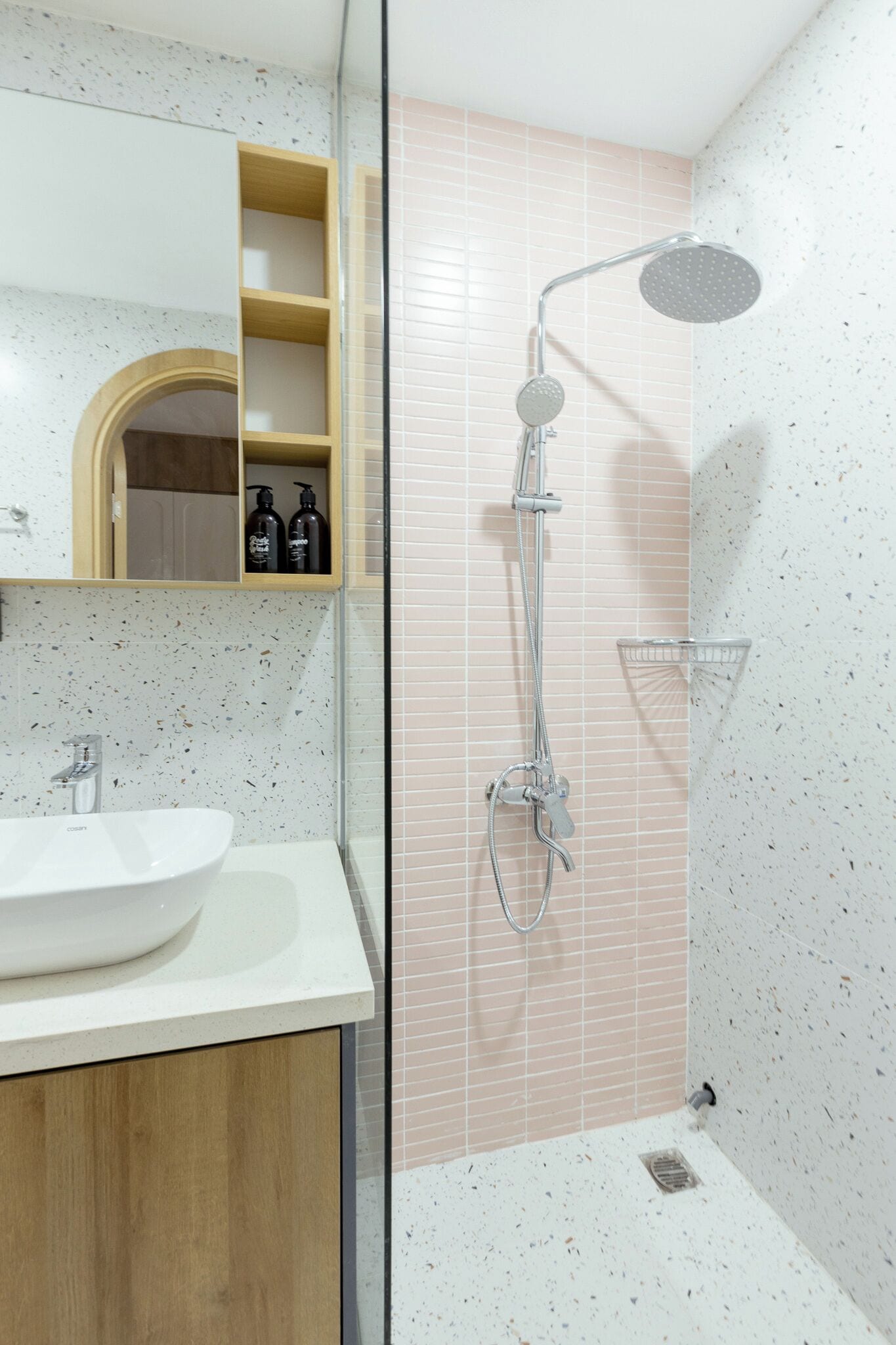 A bathroom with a white sink, a wooden cabinet, a mirror, a shower with a handheld showerhead, and a shelf with toiletries.