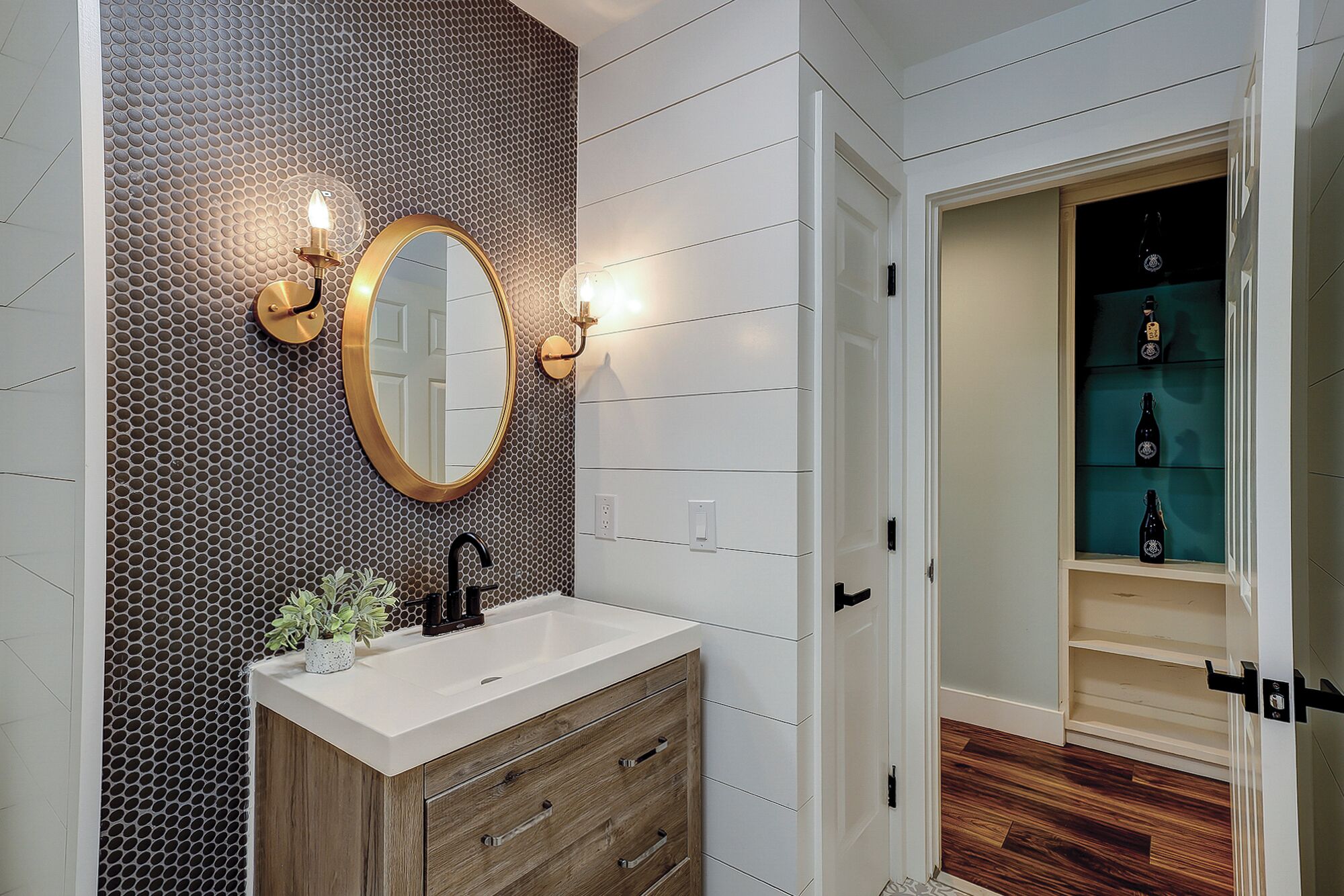 Une salle de bain avec un meuble-lavabo blanc, un miroir rond à cadre doré et un mur orné d’un motif en nid-d’abeille. On aperçoit une étagère avec des bouteilles au fond.