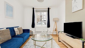 Un salon moderne avec un canapé bleu, une table basse en verre, un téléviseur posé sur un meuble en bois et une fenêtre avec des rideaux.