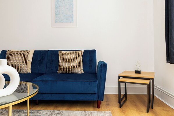 A blue sofa with patterned cushions, a small wooden table, and a glass coffee table with a white vase.