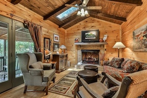 A cozy log cabin interior with a fireplace, a television mounted above it, and a comfortable seating area. - Dragonfly on the Cartecay by Mountain Oasis | Riverfront Cabin in Ellijay (Ellijay)