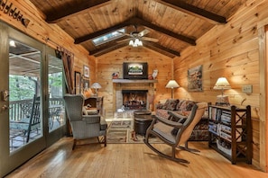 A cozy log cabin interior with a fireplace, a sofa, a rocking chair, and a bookshelf. - Dragonfly on the Cartecay by Mountain Oasis | Riverfront Cabin in Ellijay (Ellijay)