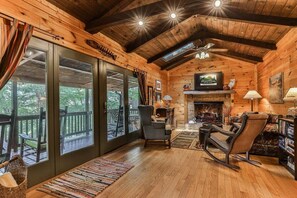 A log cabin interior with a fireplace, wooden floors, and large windows. - Dragonfly on the Cartecay by Mountain Oasis | Riverfront Cabin in Ellijay (Ellijay)