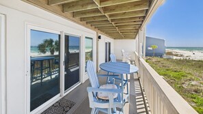 Relax in a shaded outdoor space with a dining table and chairs, overlooking the beach and ocean through large glass doors.