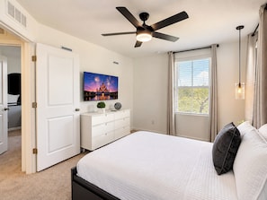 A bedroom with a bed, a ceiling fan, a window with curtains, a dresser, and a wall-mounted TV.