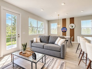 A modern living room with a grey sofa, a glass coffee table, and a dining area with chairs.