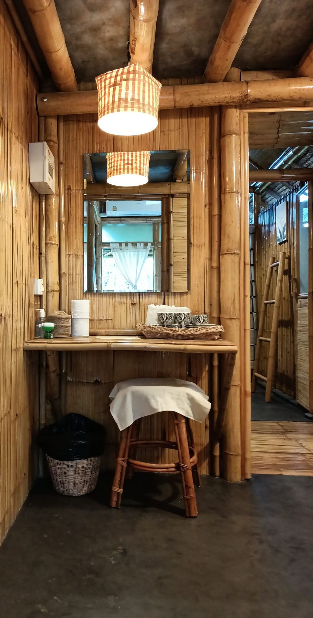 A room with bamboo walls, a wooden table with a white tablecloth, a wicker basket, a stool, and a hanging light fixture.