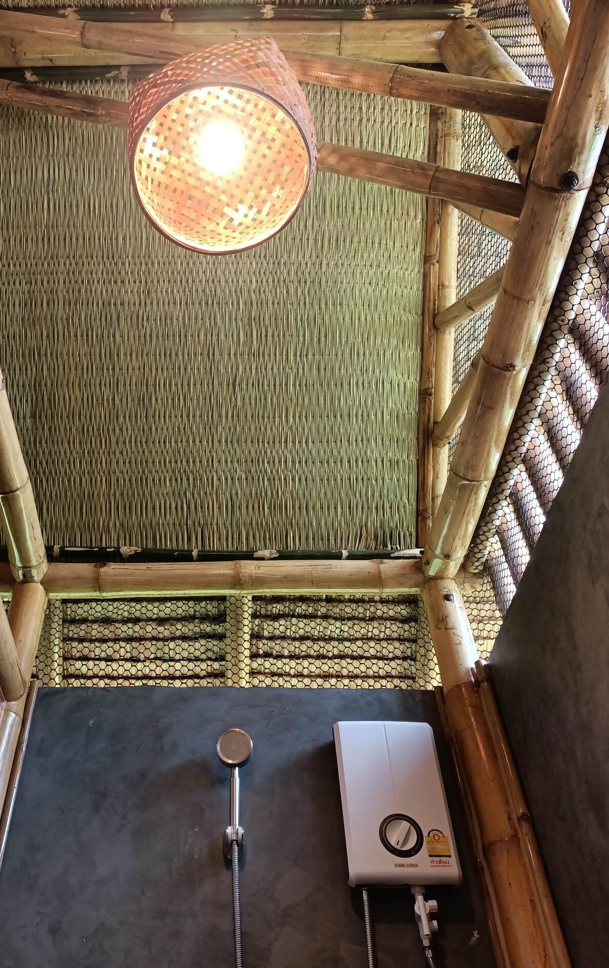 A bamboo-framed room with a woven ceiling light, a shower area, and a water heater.