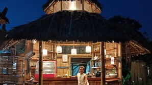 Una estructura de bar de fusta amb teulada de palla, tamborets i una persona asseguda al bar.