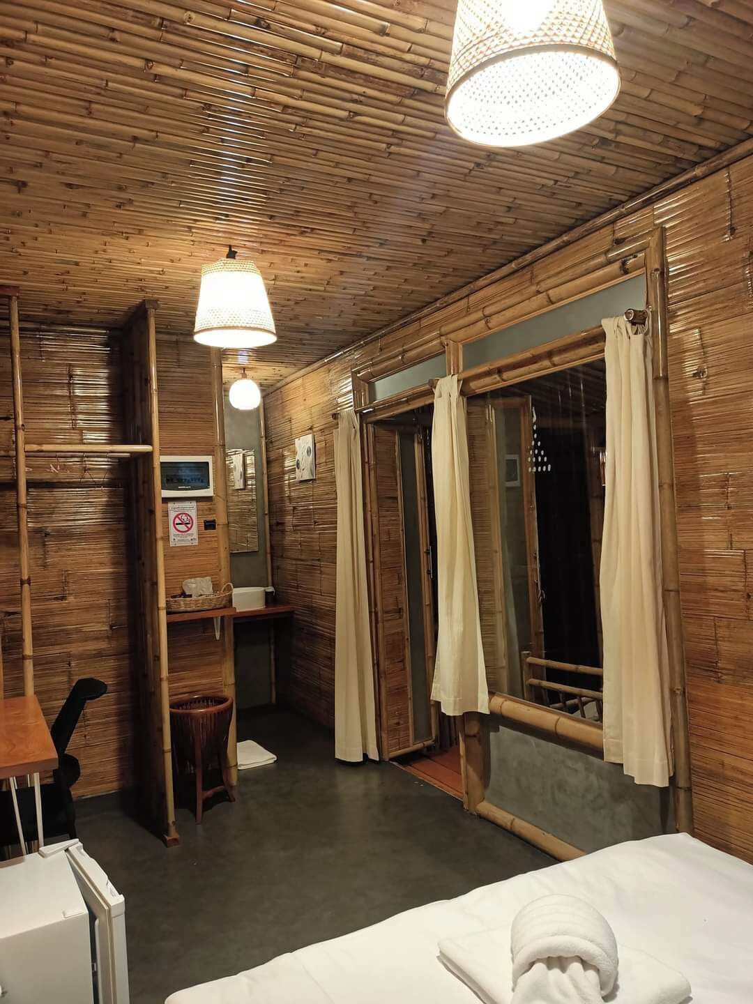 A room with wooden walls and ceiling, a bed with a white duvet, a small desk with a chair, a mirror, and a window with curtains.